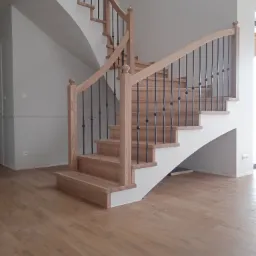 Drewniane schody z balustradą z czarnymi, kutymi elementami, prowadzące na górę, z widoczną podłogą z paneli w odcieniach drewna i szare ściany w tle.