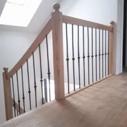 Jasne drewniane schody z czarnymi, metalowymi balustradami w nowoczesnym wnętrzu, oświetlone przez okno dachowe.