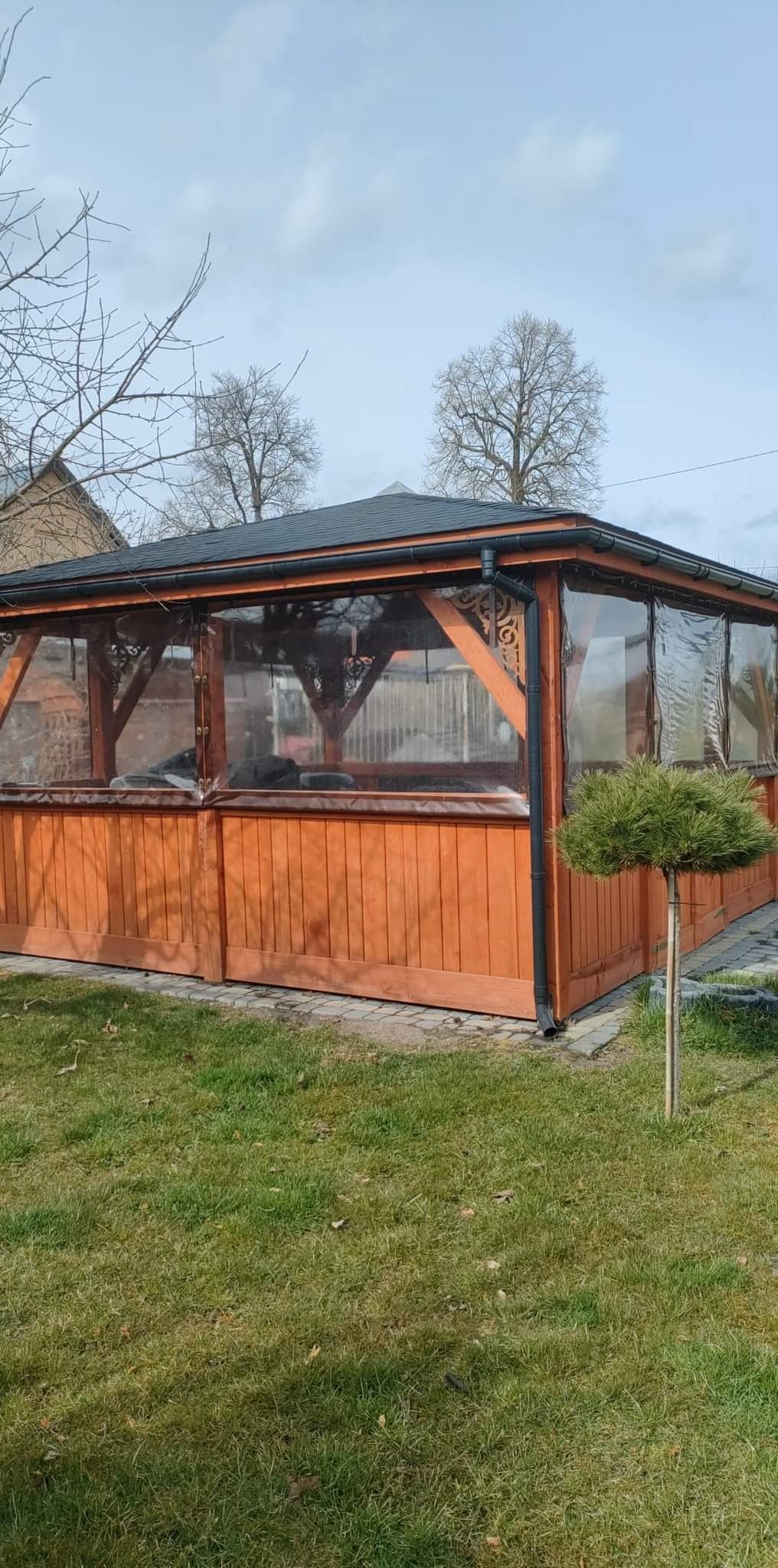 Drewniana altana z przezroczystymi ścianami, widoczna z zewnątrz, otoczona trawnikiem i młodym drzewkiem