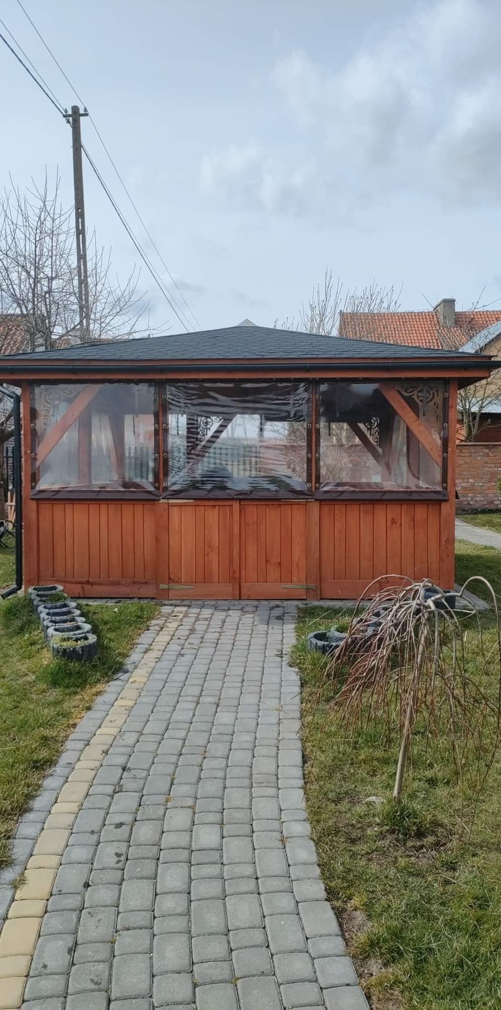 Drewniana konstrukcja z przezroczystymi ścianami osłaniającymi wnętrze, otoczona trawnikiem i ścieżką z kostki brukowej, z widocznymi elementami dekoracyjnymi z opon.