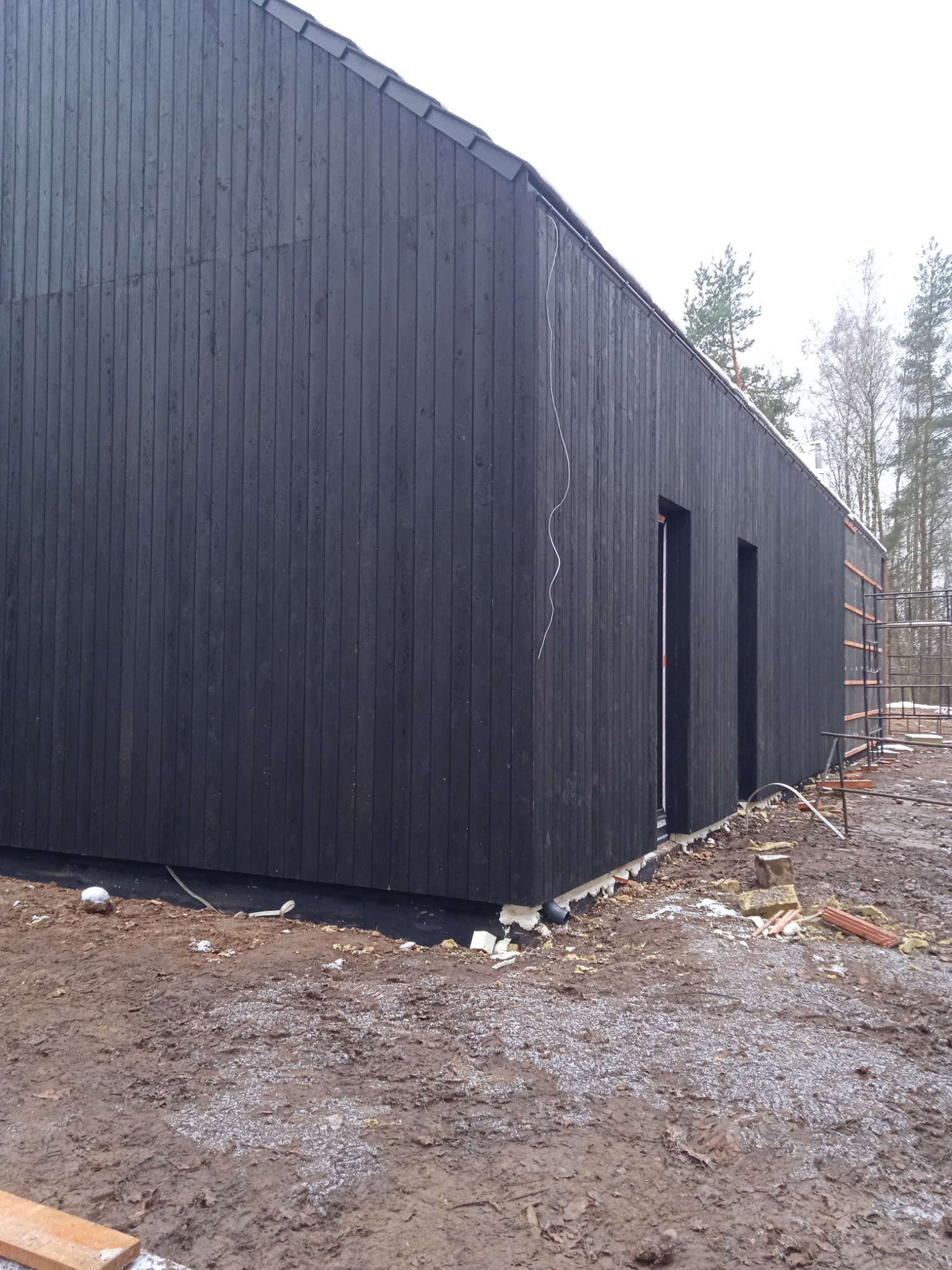 Ciemna, drewniana elewacja budynku w trakcie budowy, widoczne fragmenty izolacji i rusztowania, teren wokół budynku częściowo pokryty błotem.