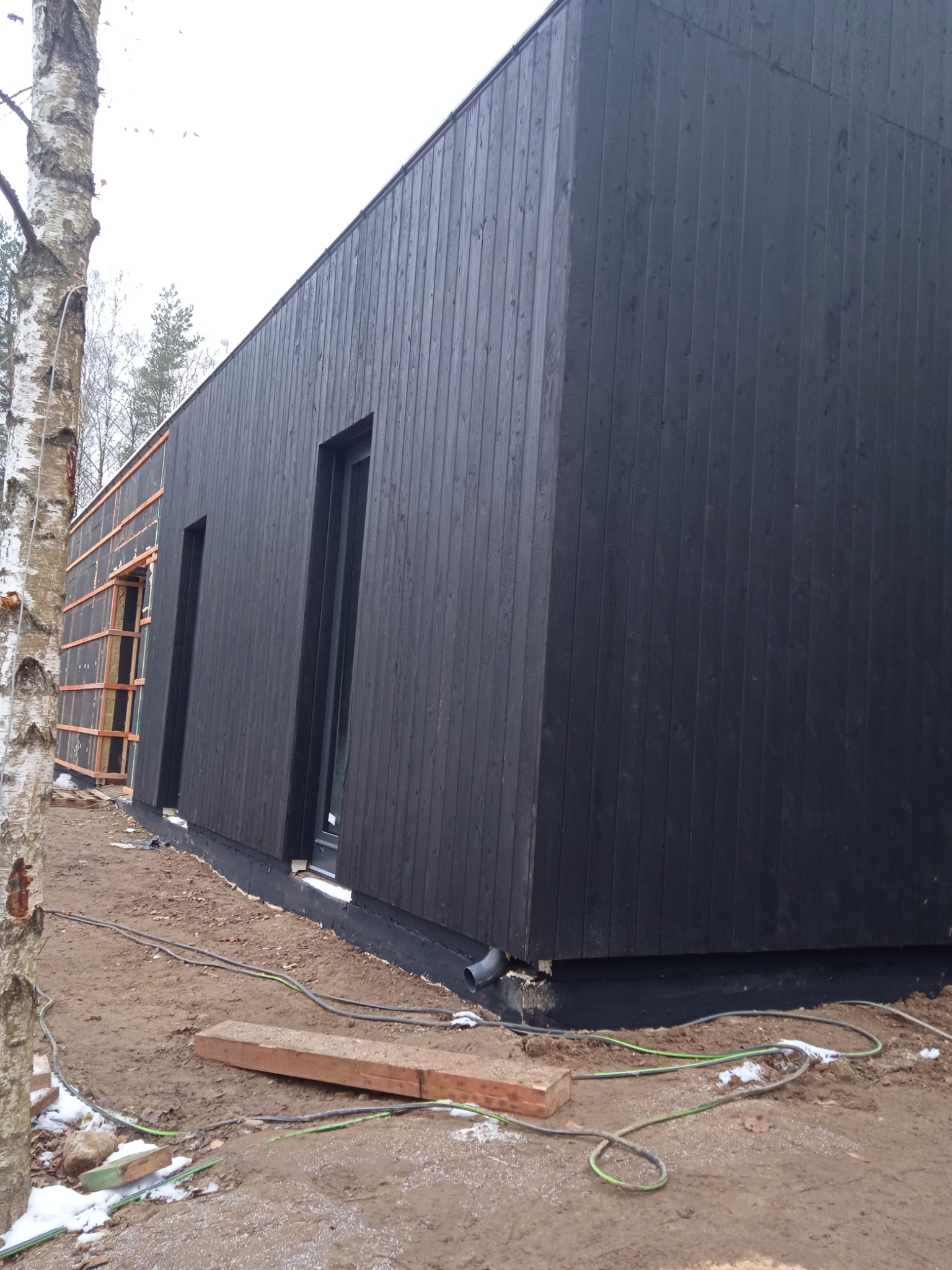 Ciemna, drewniana elewacja budynku w trakcie budowy, widoczne okna i fragment ocieplenia, teren wokół w stanie surowym.