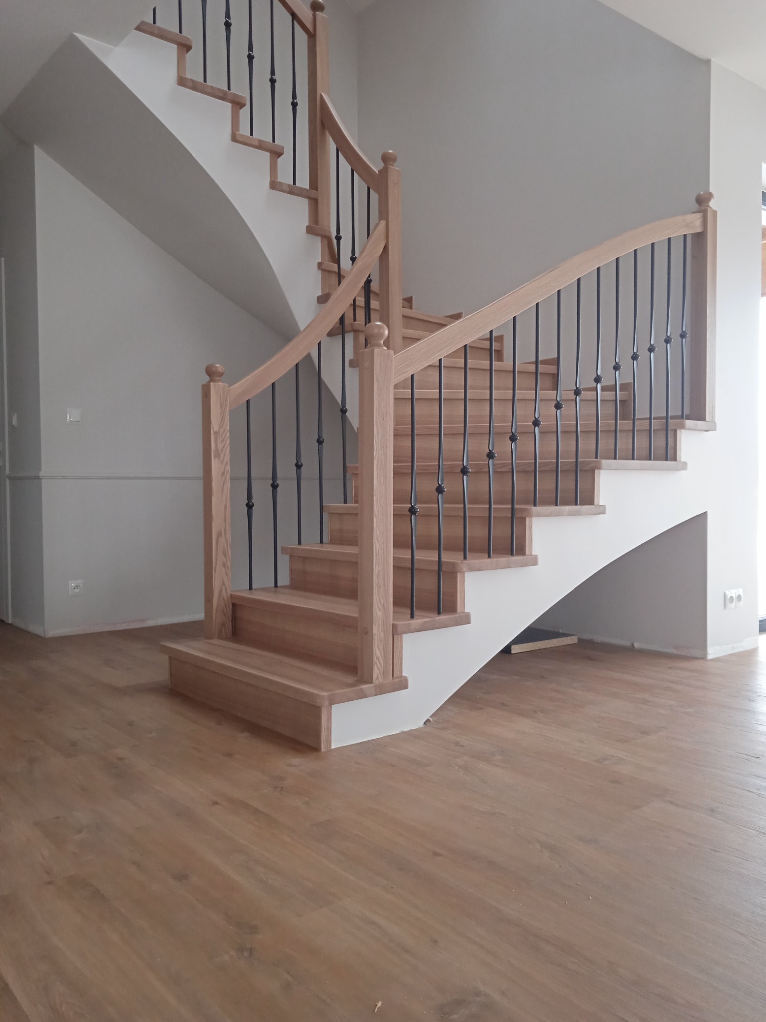 Drewniane schody z balustradą z czarnymi, kutymi elementami, prowadzące na górę, z widoczną podłogą z paneli w odcieniach drewna i szare ściany w tle.