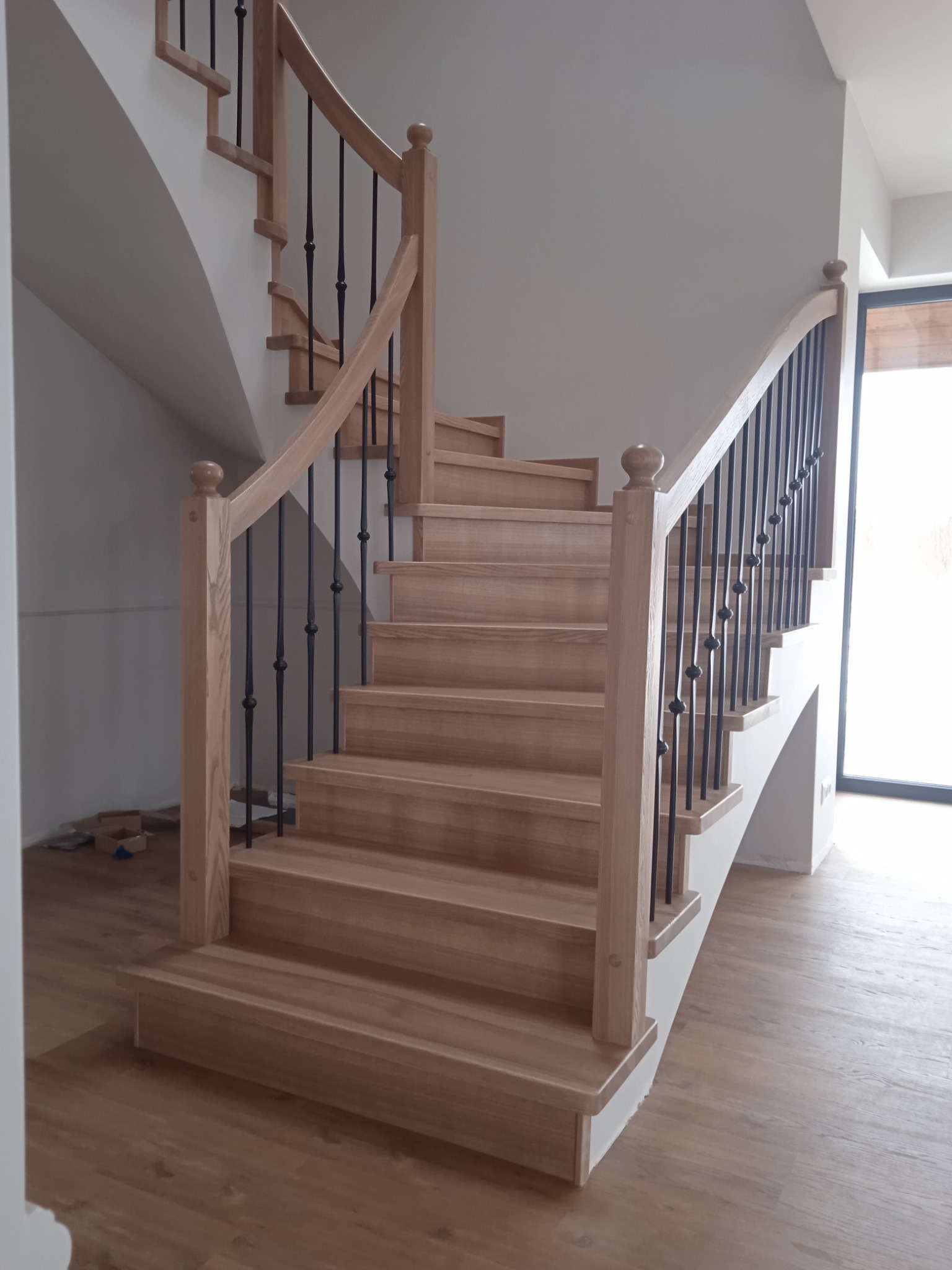 Jasne, drewniane schody z czarnymi, kutymi balustradami w nowoczesnym wnętrzu. Widoczne drewniane stopnie i poręcze z okrągłymi zakończeniami. Podłoga wyłożona panelami imitującymi jasne drewno.