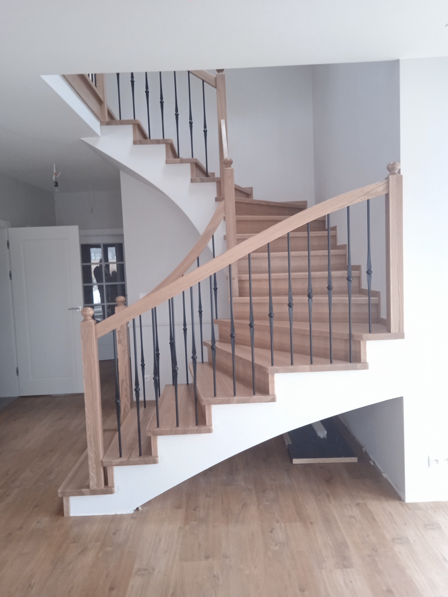 Spiralne schody z drewna z czarnymi, metalowymi balustradami, prowadzące na wyższe piętro, z widocznymi elementami wykończeniowymi i jasną podłogą.