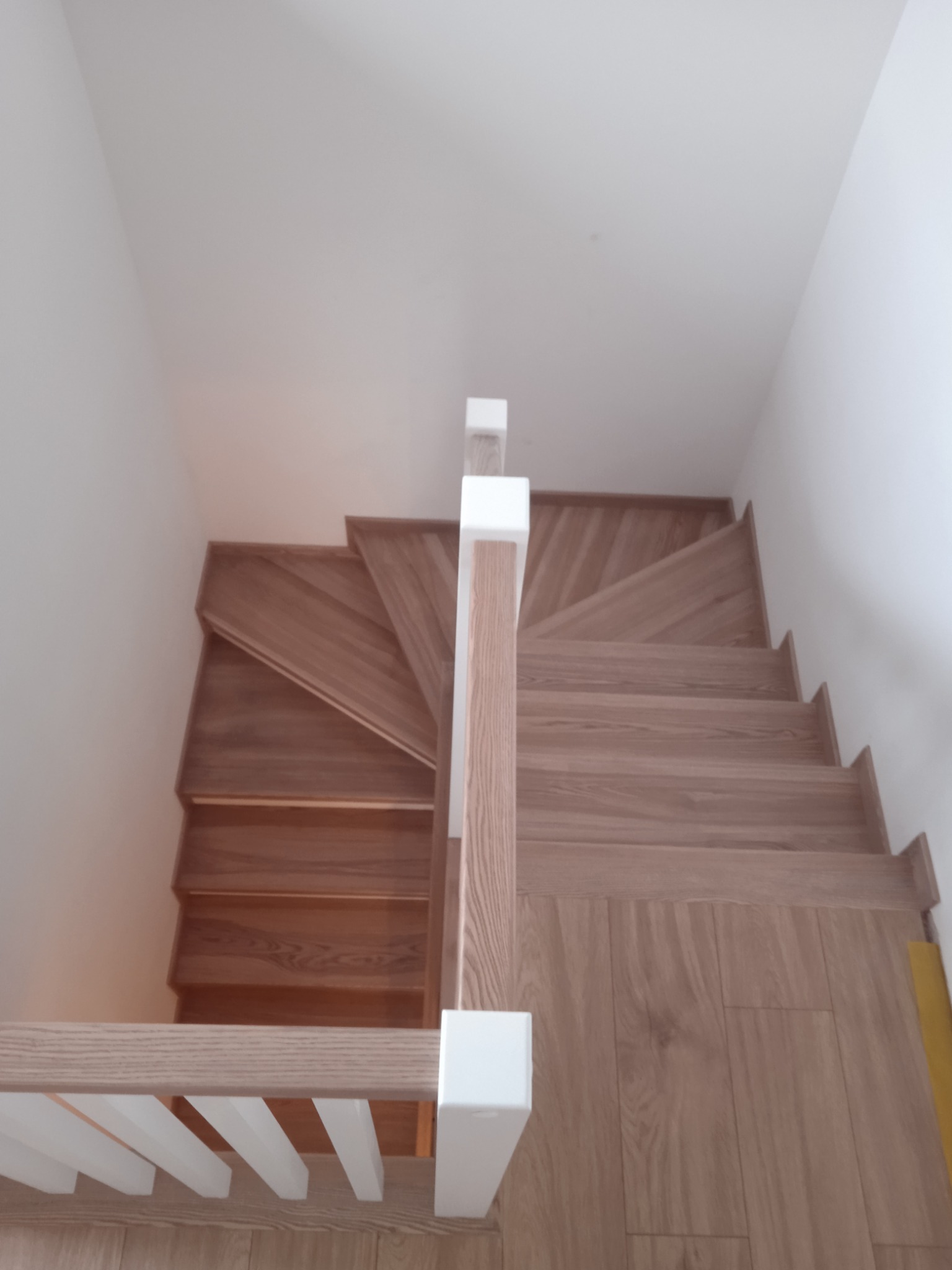 Drewniane schody z białymi balustradami, widok z góry, ukazujący zakręt schodów i naturalne usłojenie drewna.