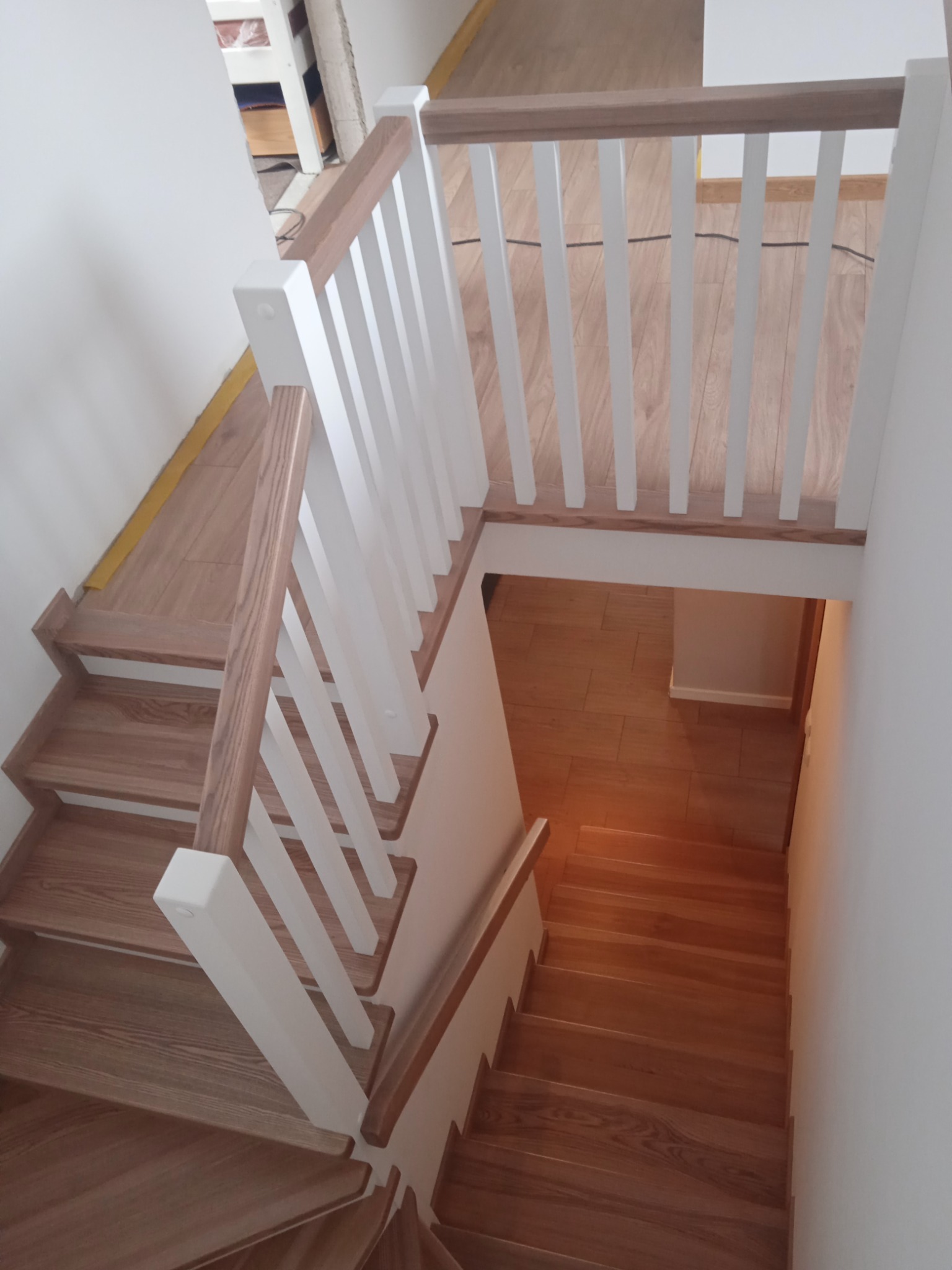 Widok z góry na drewniane schody z białą balustradą, jasne drewno stopni kontrastuje z białymi elementami balustrady, widoczne niedokończone wykończenia na górze schodów.