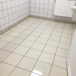 Błyszcząca, jasna podłoga z kwadratowych płytek w czystej łazience biurowej, widoczna umywalka i grzejnik na ścianie z białych płytek.