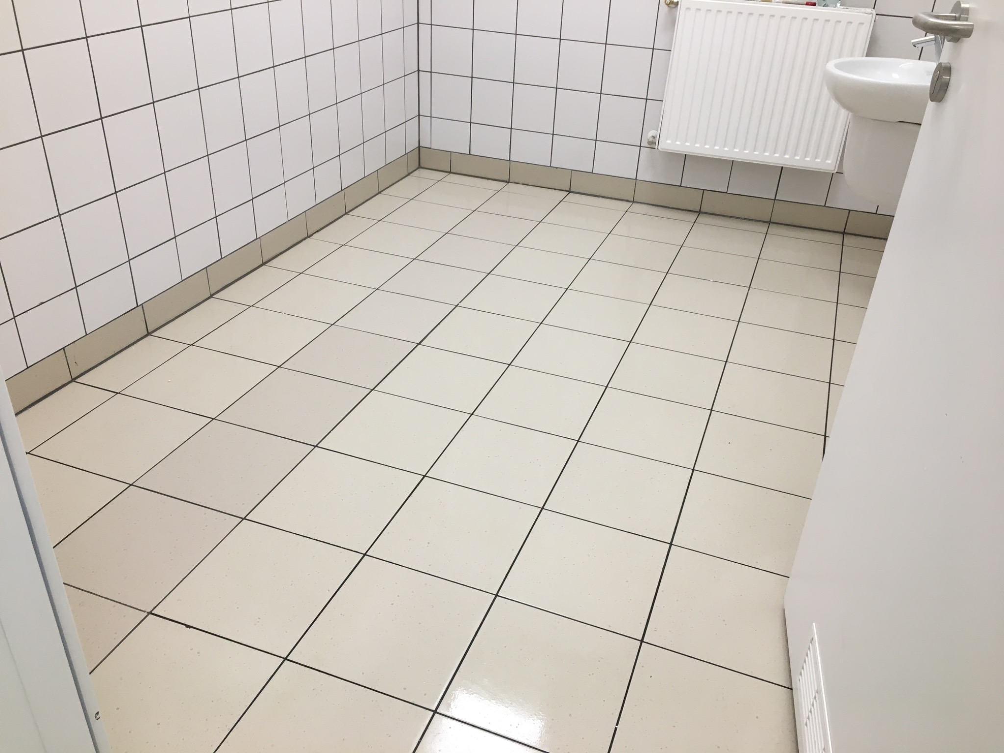 Błyszcząca, jasna podłoga z kwadratowych płytek w czystej łazience biurowej, widoczna umywalka i grzejnik na ścianie z białych płytek.