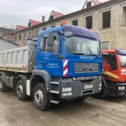 Dwa ciężarowe samochody MAN: niebieski wywrotka i czerwony z napisem 'Odpady', stojące na placu z budynkami w tle, widoczne tablice rejestracyjne.