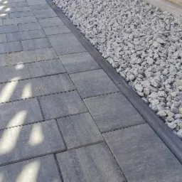 Szara kostka brukowa ułożona w Tomaszowie Mazowieckim, obok ozdobny pas z białego grysu, całość oświetlona promieniami słońca, rzucającymi cienie drzew.