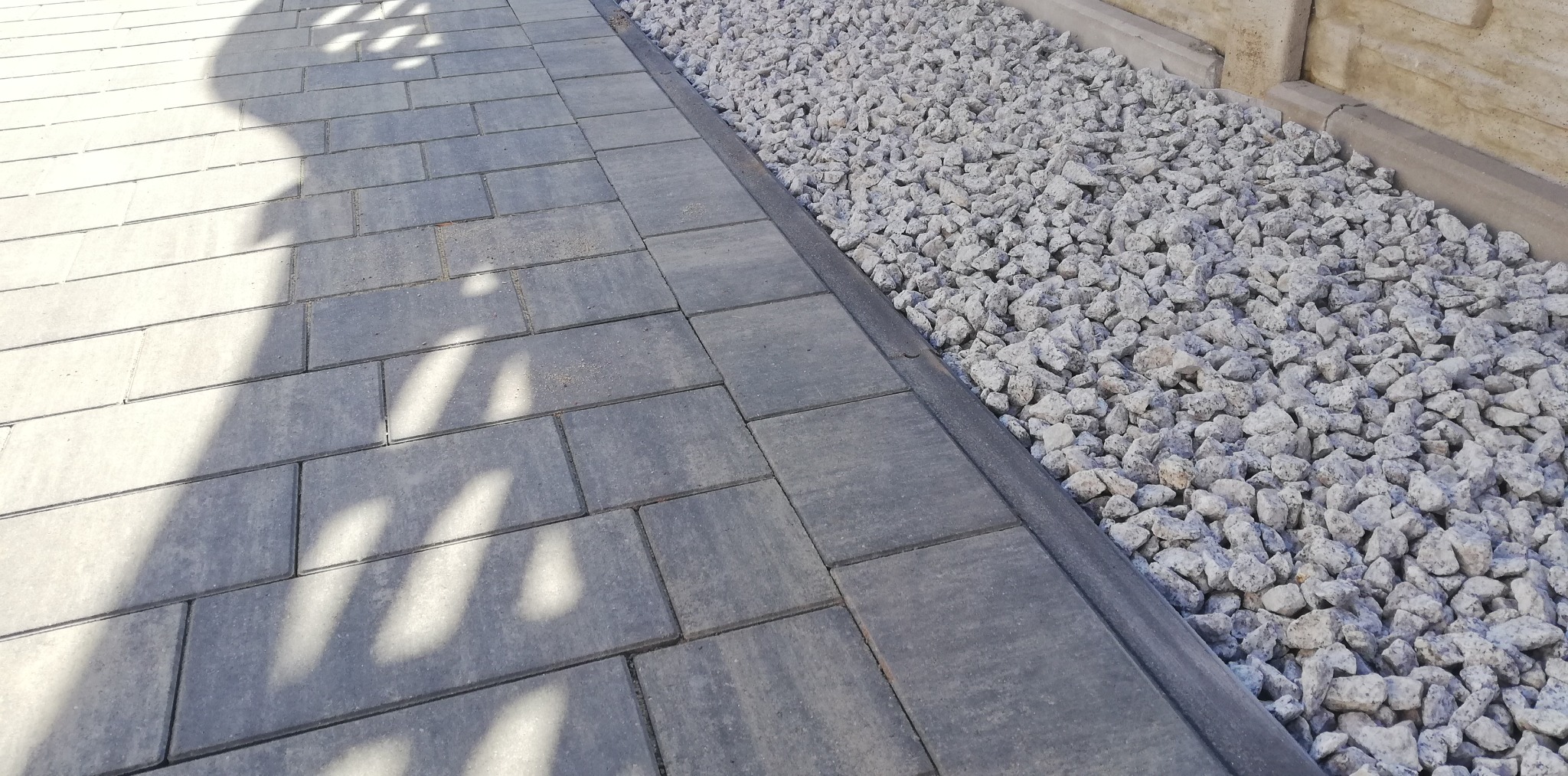 Szara kostka brukowa ułożona w Tomaszowie Mazowieckim, obok ozdobny pas z białego grysu, całość oświetlona promieniami słońca, rzucającymi cienie drzew.