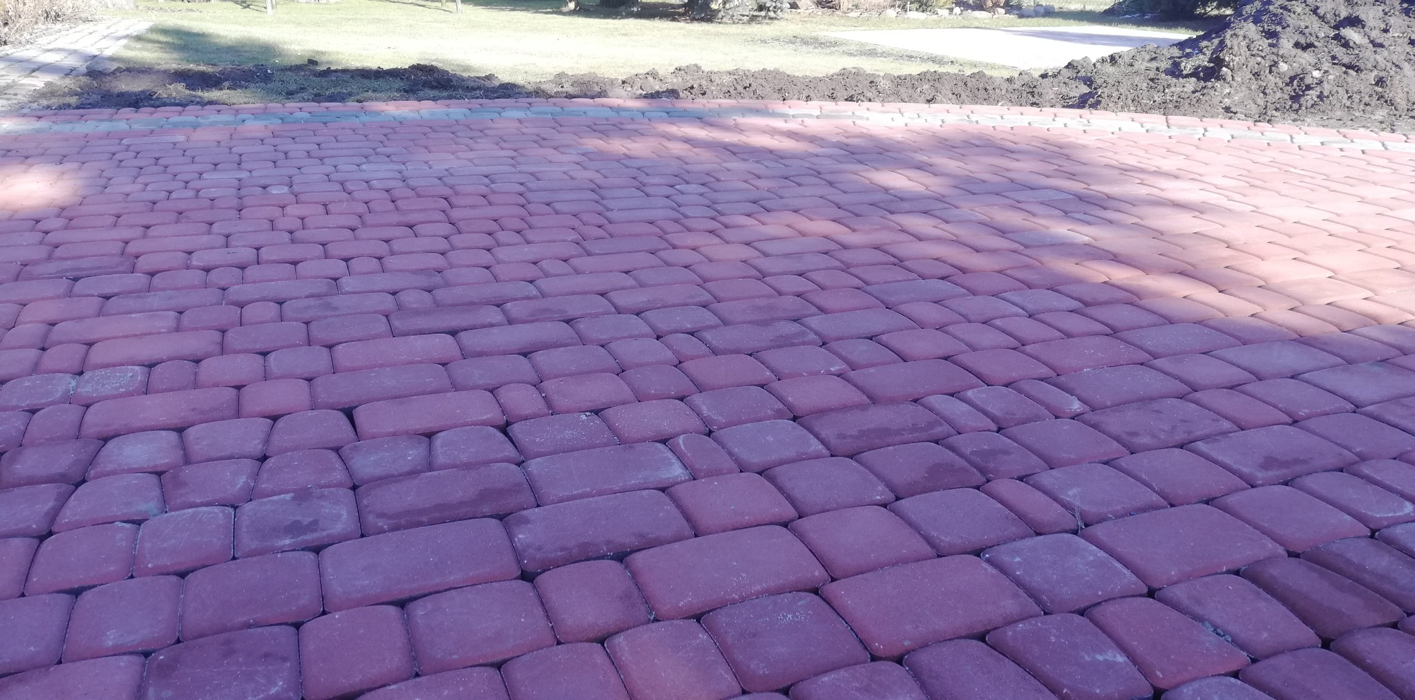 Ułożona, czerwona kostka brukowa w regularnym wzorze, z widoczną linią oddzielającą od przygotowanego terenu pod dalsze prace na trawniku w tle.