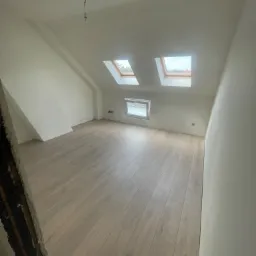 Poddasze z dwiema oknami dachowymi i jasną podłogą z paneli, świeżo pomalowane ściany na biało, widoczny fragment ościeżnicy w lewym dolnym rogu.