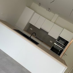 EasyFinish - Nowoczesna, minimalistyczna kuchnia w Brzegu z białymi szafkami, drewnianym blatem i czarną armaturą. Widoczne niedokończone podłączenia elektryczne sugerują etap wykończenia.