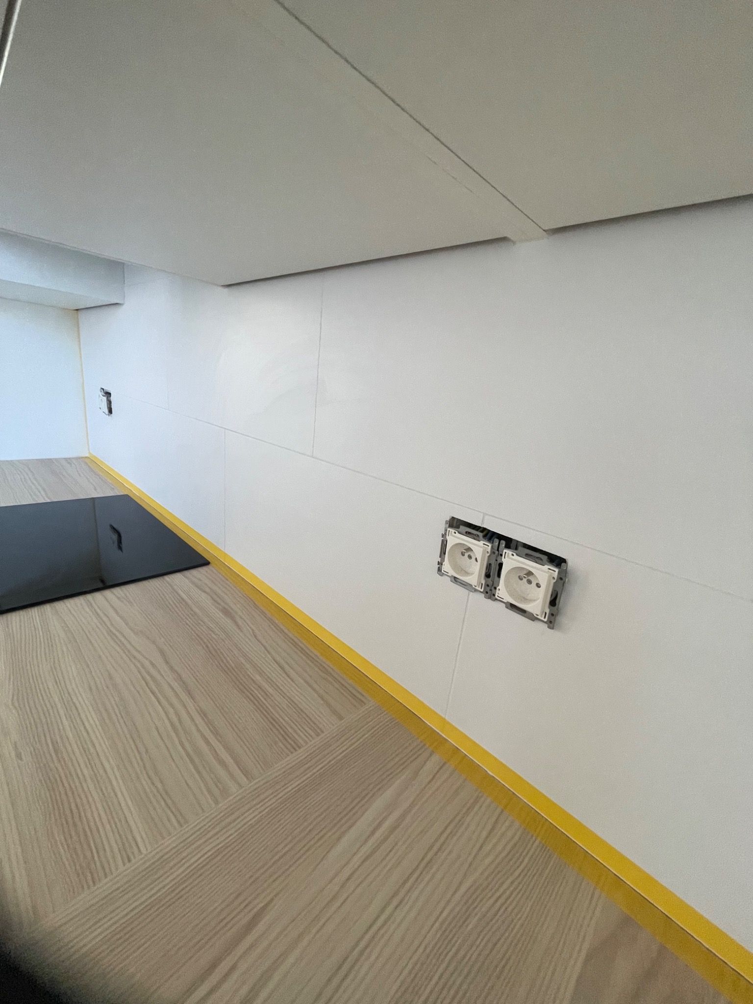 Fragment kuchni z białymi płytkami ściennymi, drewnopodobnymi panelami podłogowymi i podwójnym gniazdkiem elektrycznym bez osprzętu, zabezpieczonym żółtą taśmą malarską.