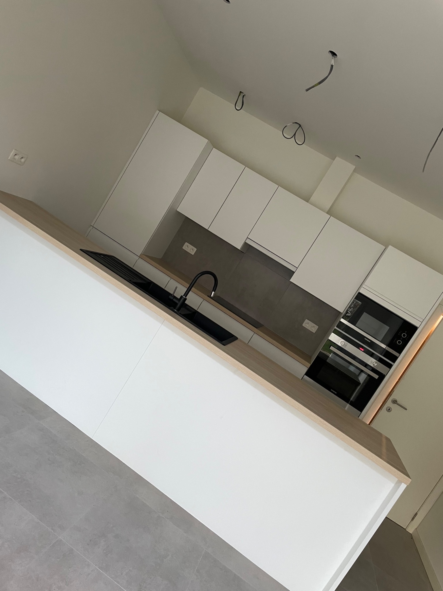 Nowoczesna, minimalistyczna kuchnia w Brzegu z białymi szafkami, drewnianym blatem i czarną armaturą. Widoczne niedokończone podłączenia elektryczne sugerują etap wykończenia.