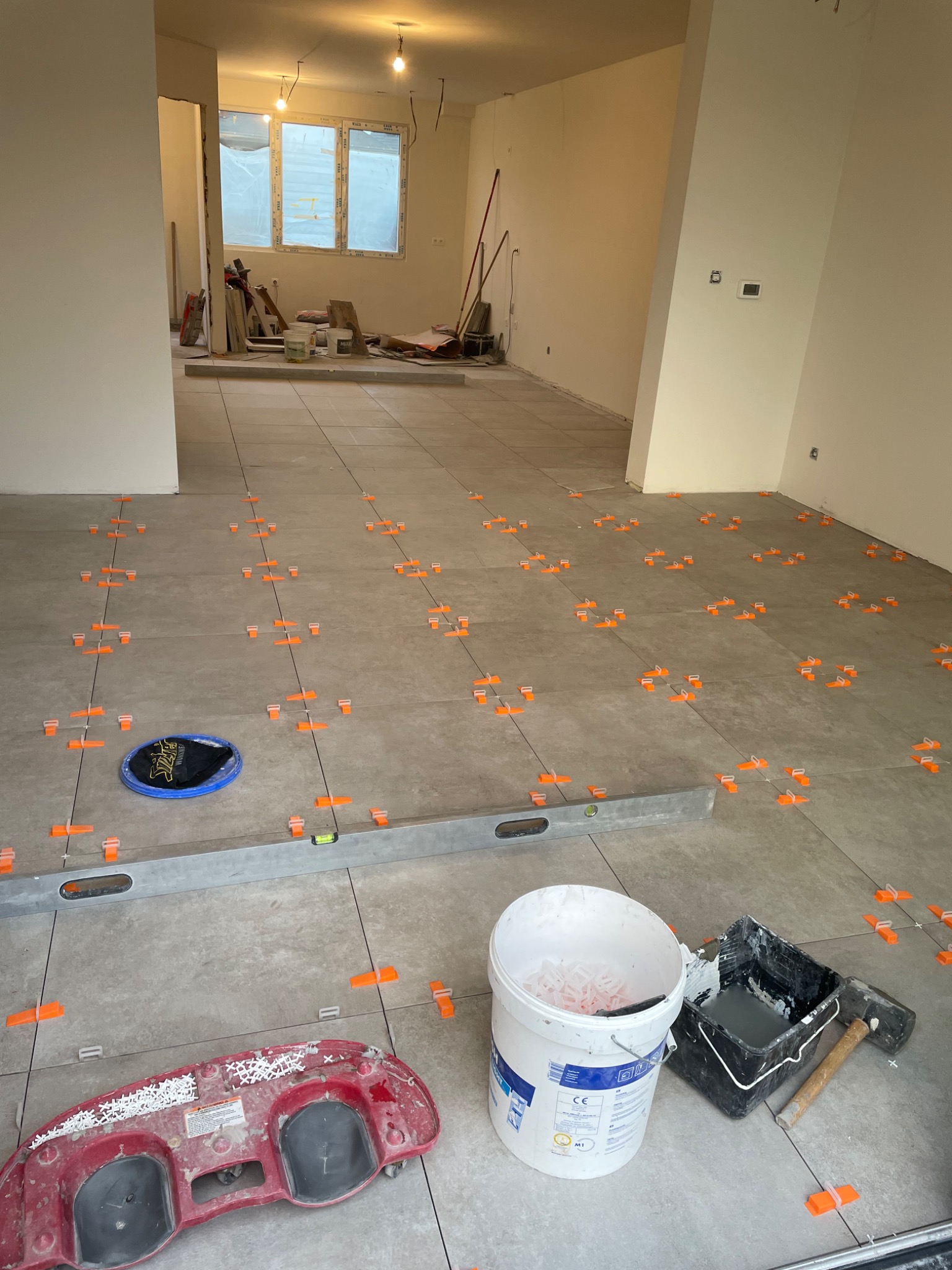 Szare płytki podłogowe w trakcie układania, z pomarańczowymi klipsami poziomującymi, poziomica i narzędzia glazurnicze w nieukończonym pomieszczeniu.