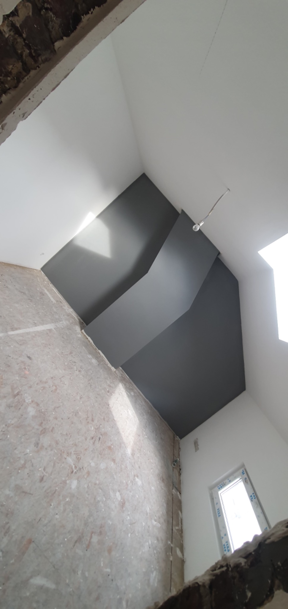 Wnętrze w trakcie remontu: surowa ściana z widocznymi śladami po instalacjach, pomalowany na szaro fragment sufitu z nietypowym załamaniem, biała ściana z oknem w tle.