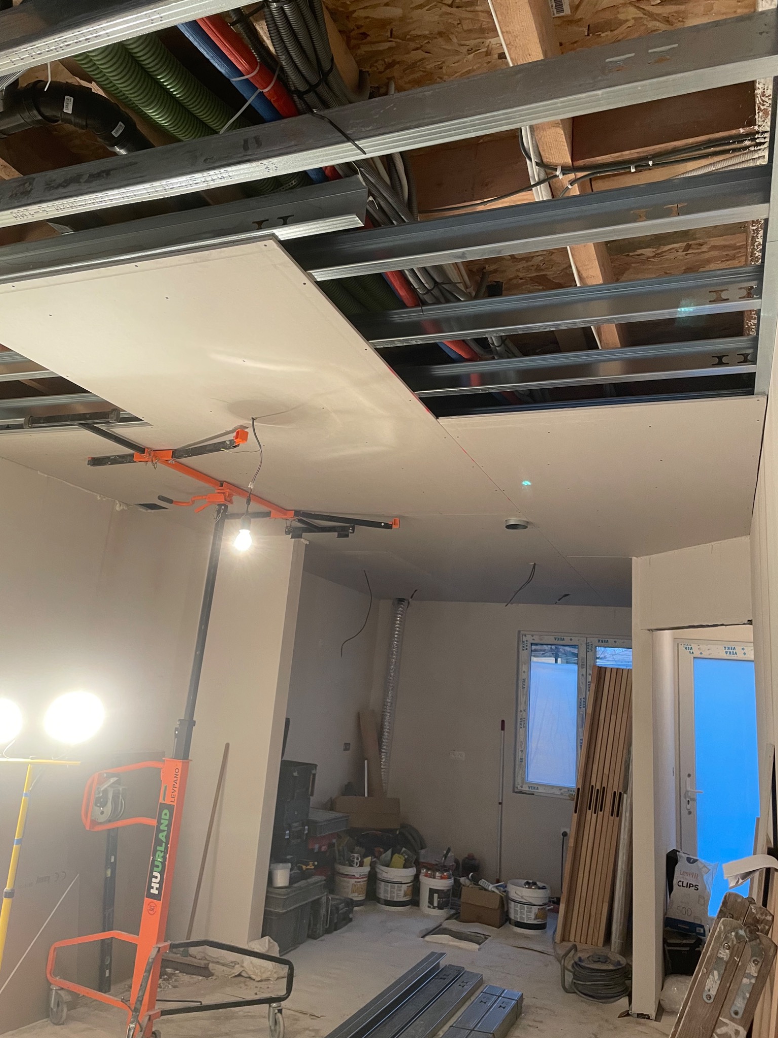 Montaż sufitu podwieszanego z płyt gipsowo-kartonowych, widoczny stelaż metalowy, podnośnik do płyt Huurland Levpano i instalacje pod sufitem.