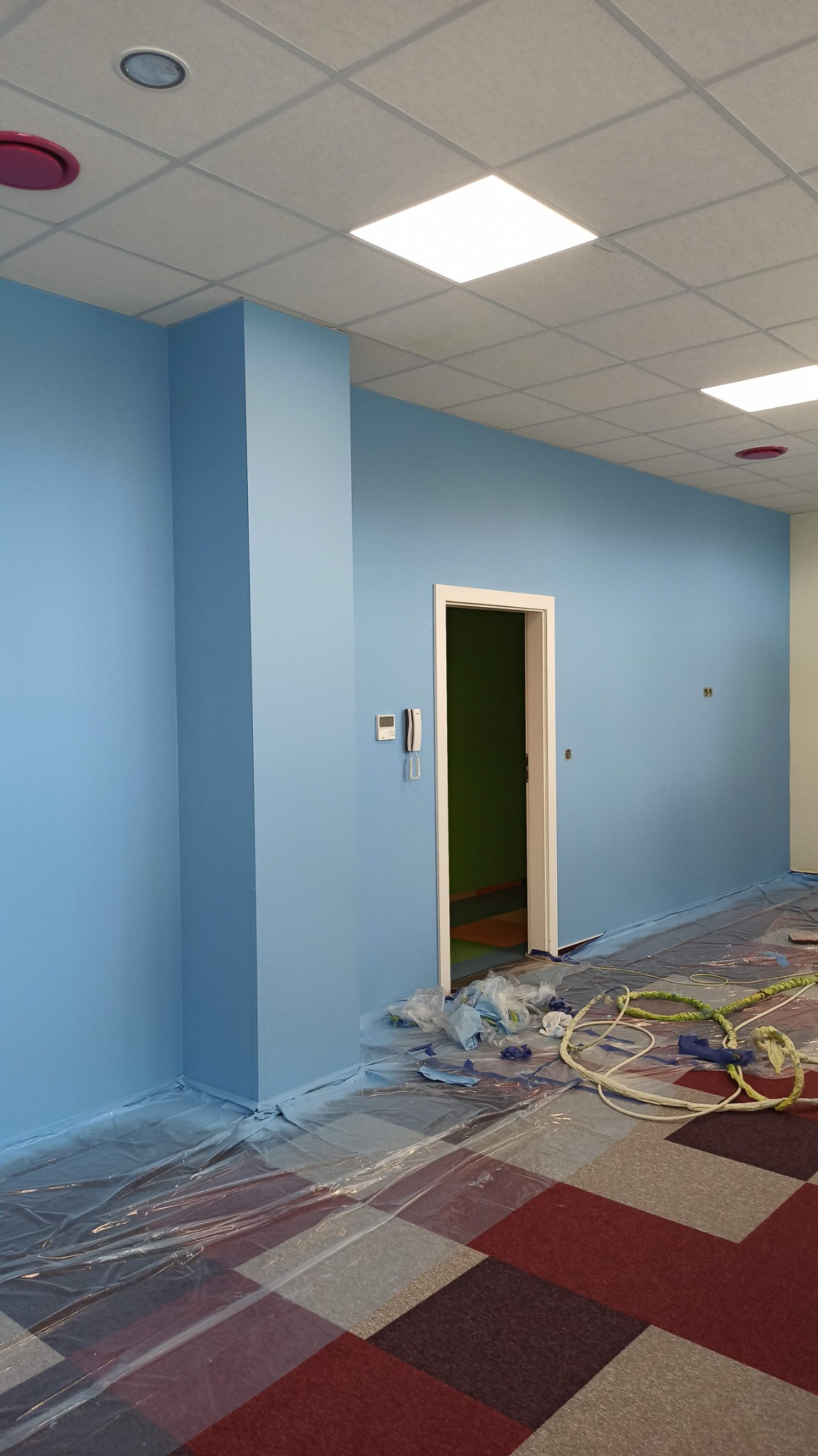 Świeżo pomalowane na błękitno ściany biura z widocznymi zabezpieczeniami podłogi folią i resztkami farby przy listwach przypodłogowych. Na ścianie widoczny telefon stacjonarny.