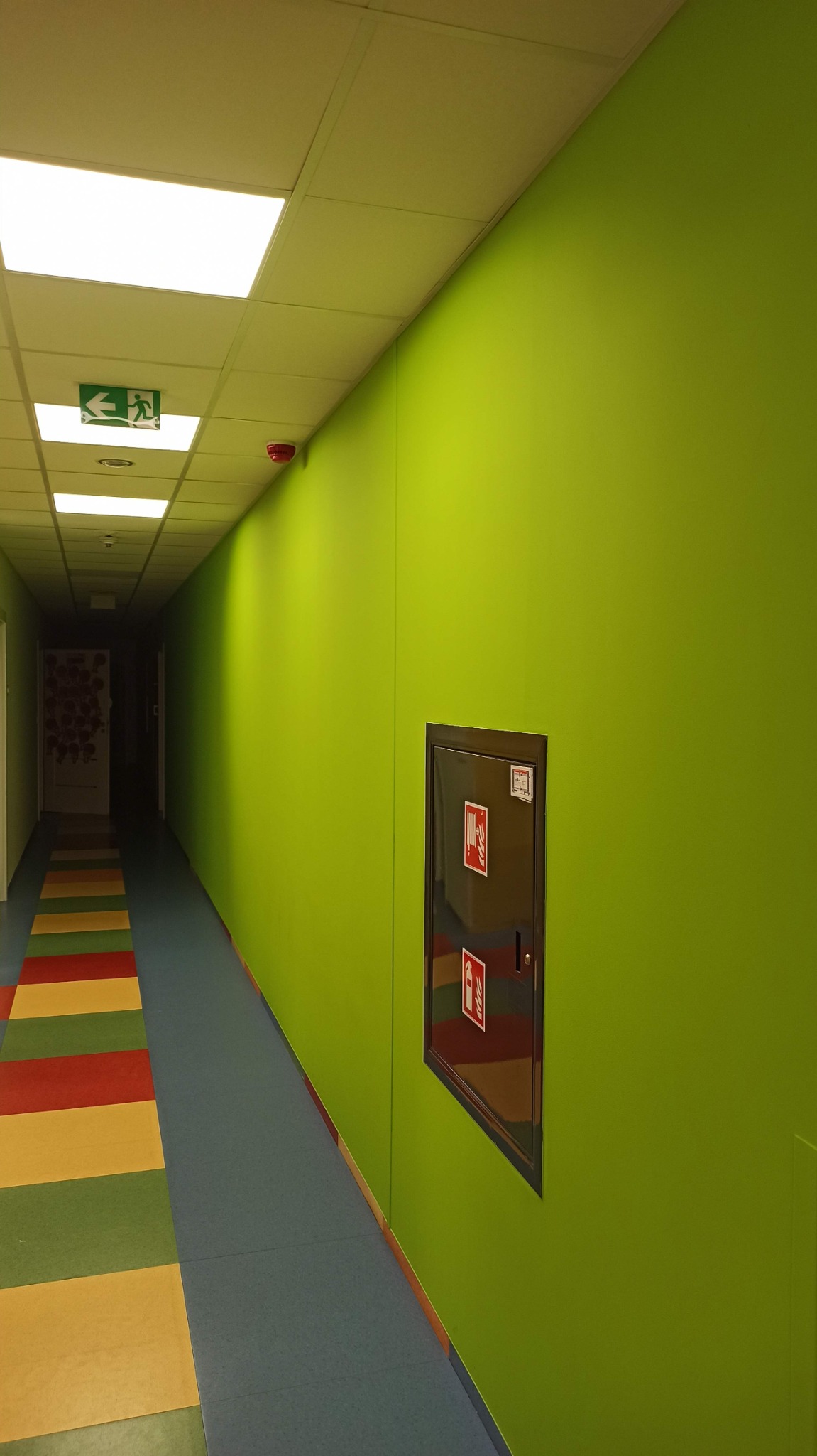 Długi korytarz biurowy z jasnozielonymi ścianami, kolorową podłogą i metalową szafką przeciwpożarową w ścianie.