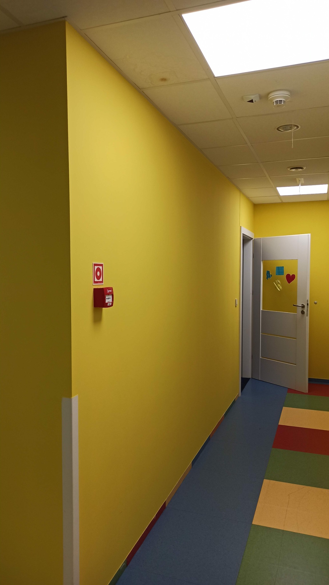 Żółty korytarz biurowy z kolorową podłogą, białą listwą przypodłogową, czujnikiem dymu na suficie i czerwoną skrzynką przeciwpożarową na ścianie.