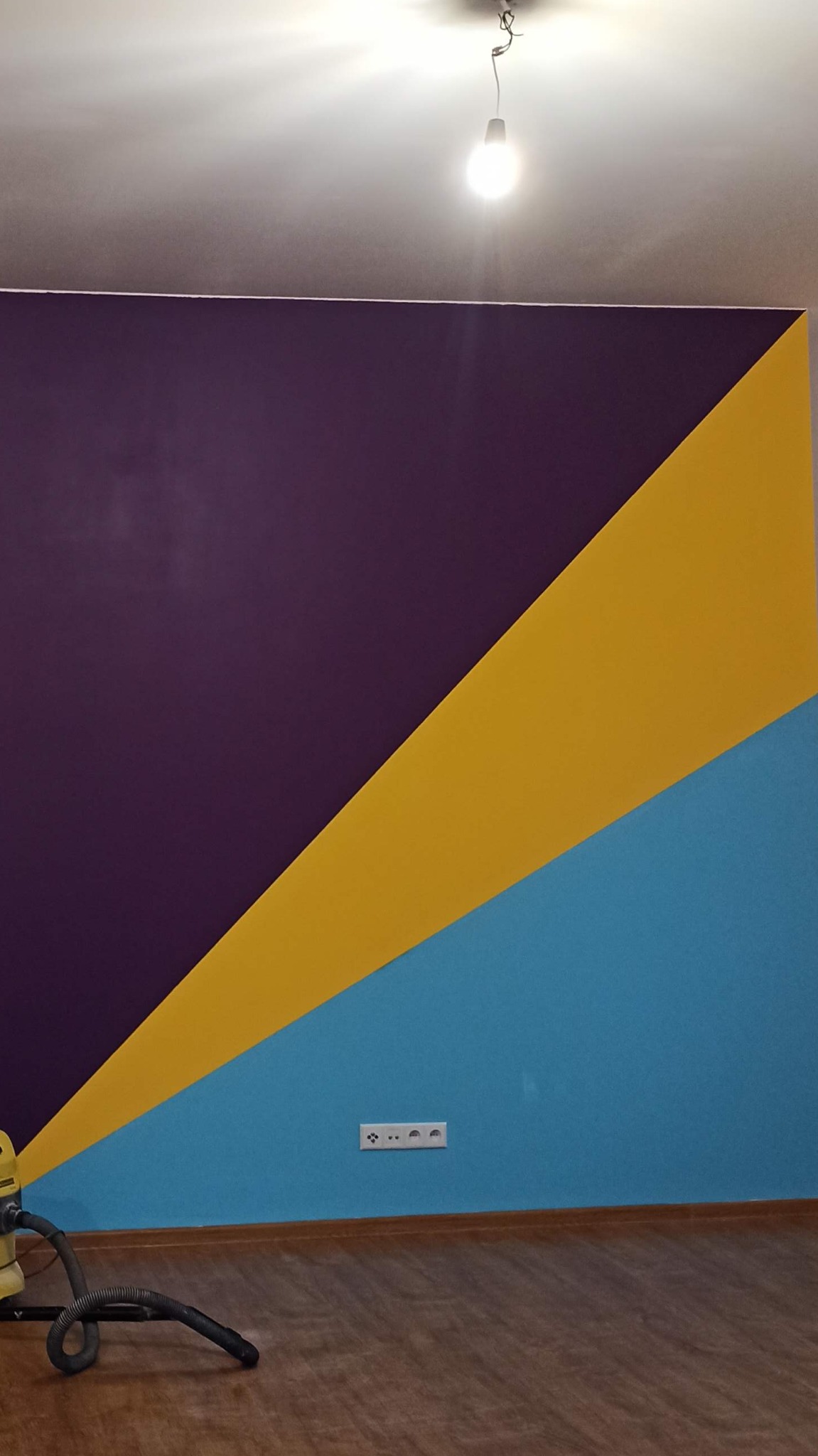 Pomieszczenie po malowaniu ścian w geometryczne wzory: fiolet, żółty i błękit, widoczny odkurzacz przy podłodze.