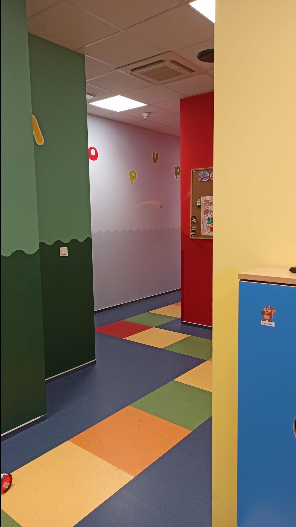 Korytarz w przedszkolu z kolorowymi ścianami i podłogą w kwadraty, na ścianach naklejone litery, na szafce naklejka z wiewiórką.