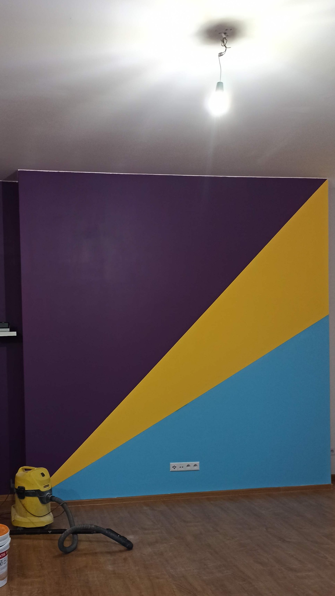 Pomieszczenie po malowaniu ściany w geometryczne wzory: fioletowy, żółty i niebieski, widoczny odkurzacz i gniazdko elektryczne.
