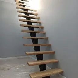 Nowoczesne schody z drewnianymi stopniami i czarną metalową konstrukcją nośną, widoczne z dołu w surowym wnętrzu z zabezpieczoną folią podłogą.