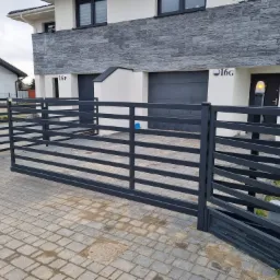 Nowoczesna, pozioma brama wjazdowa w kolorze antracytowym, zamontowana przed domem z dwoma garażami i elewacją z szarej cegły, kostka brukowa na podjeździe.