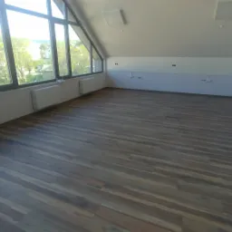 Wnętrze poddasza z ułożoną, ciemnobrązową wykładziną podłogową imitującą drewno, widok na okna dachowe z widokiem na zabudowę mieszkalną i zieleń.