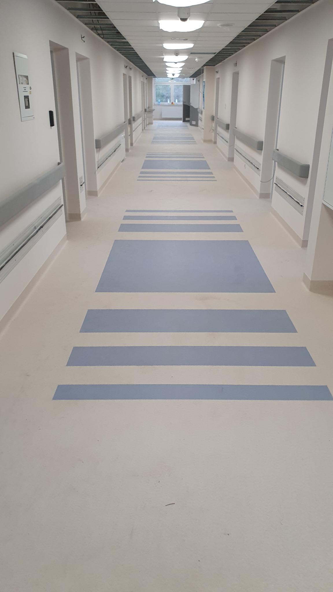Długi korytarz w placówce medycznej z jasną, beżową podłogą i niebieskimi, geometrycznymi wzorami. Na ścianach widoczne poręcze i drzwi do pomieszczeń.