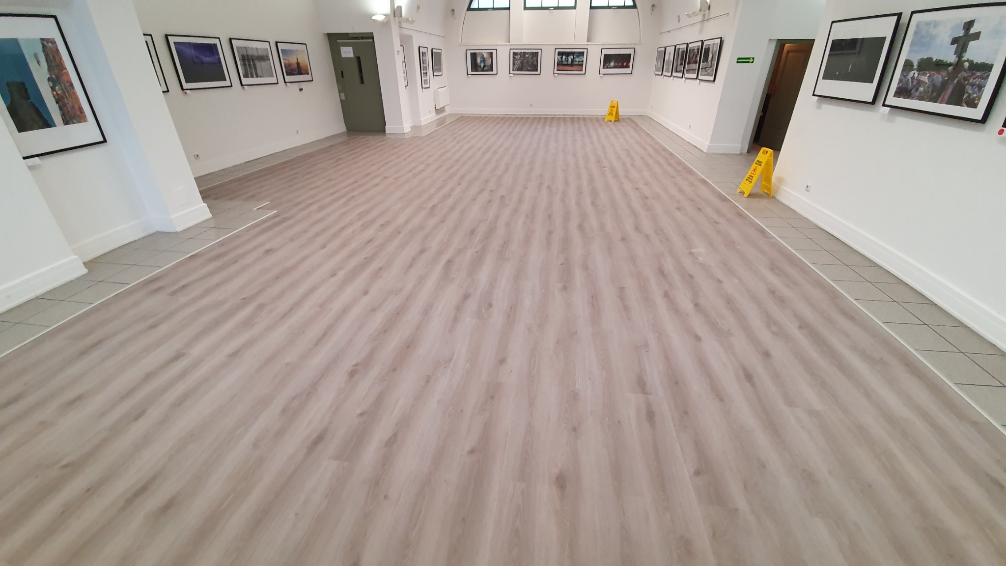 Jasna, pusta sala wystawowa z nową, drewnopodobną podłogą, zabezpieczona żółtymi znakami ostrzegawczymi przed śliską powierzchnią.