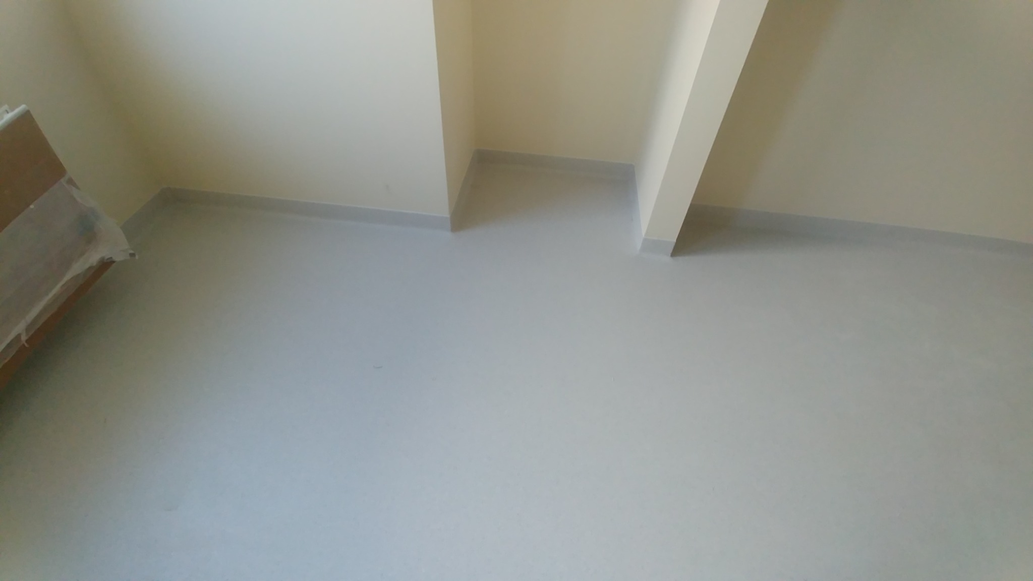 Jasnoszara, gładka podłoga w pustym pomieszczeniu z beżowymi ścianami i listwami przypodłogowymi.