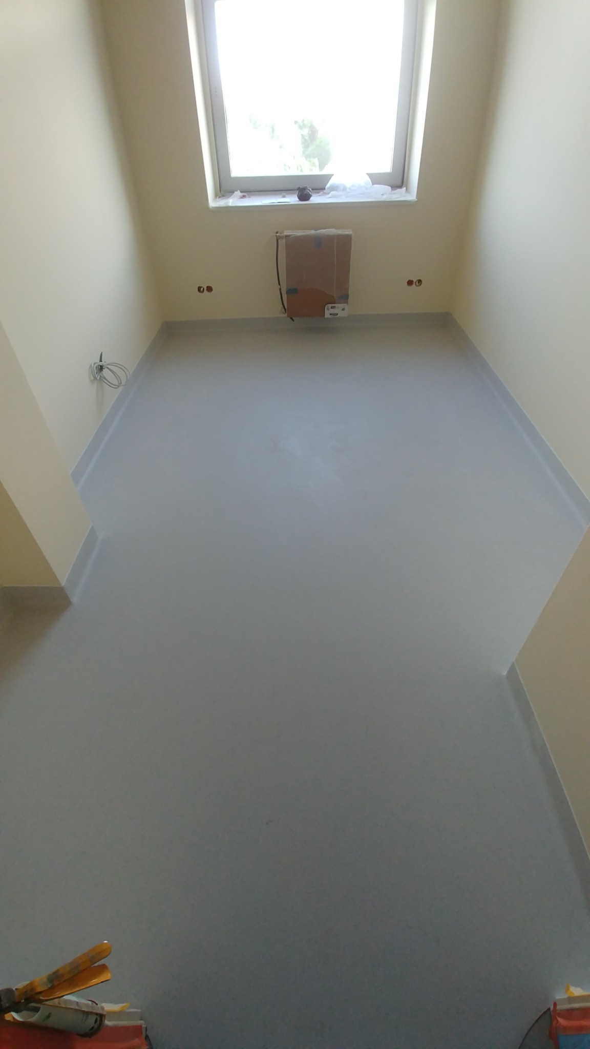 Szara, jednolita podłoga w pomieszczeniu ze skosami, widoczne listwy przypodłogowe, okno dachowe z pudełkiem pod nim, instalacja elektryczna na ścianie i narzędzia w rogu.