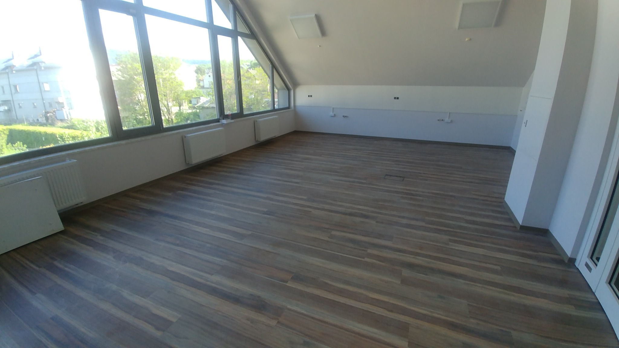 Wnętrze poddasza z ułożoną, ciemnobrązową wykładziną podłogową imitującą drewno, widok na okna dachowe z widokiem na zabudowę mieszkalną i zieleń.