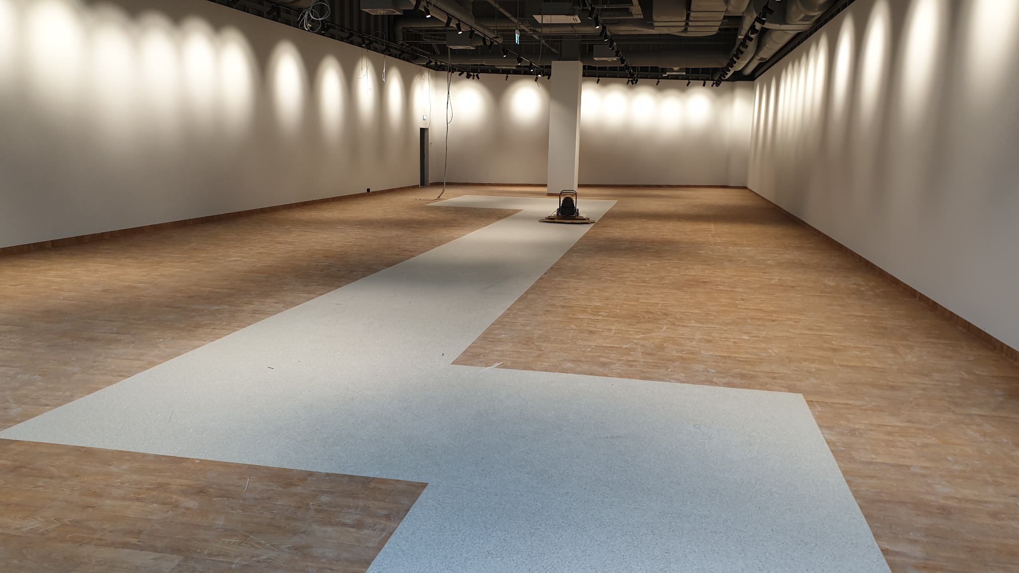 Wnętrze dużego pomieszczenia z częściowo ułożoną jasną podłogą, widoczna maszyna do polerowania podłóg i surowe ściany z oświetleniem punktowym.