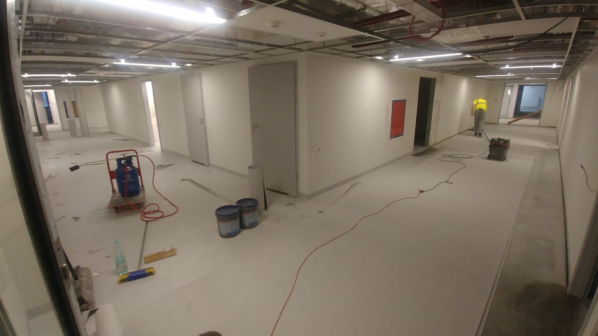 Rozległy korytarz w trakcie prac wykończeniowych: świeżo położona, jasna wykładzina, butla gazowa z palnikiem na wózku, puszki farby i pracownik w oddali z odkurzaczem przemysłowym.