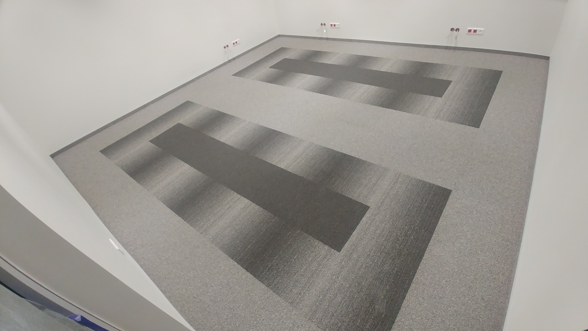 Szara wykładzina dywanowa ułożona w geometryczny wzór w pustym pomieszczeniu biurowym, widoczne gniazdka elektryczne na ścianie.