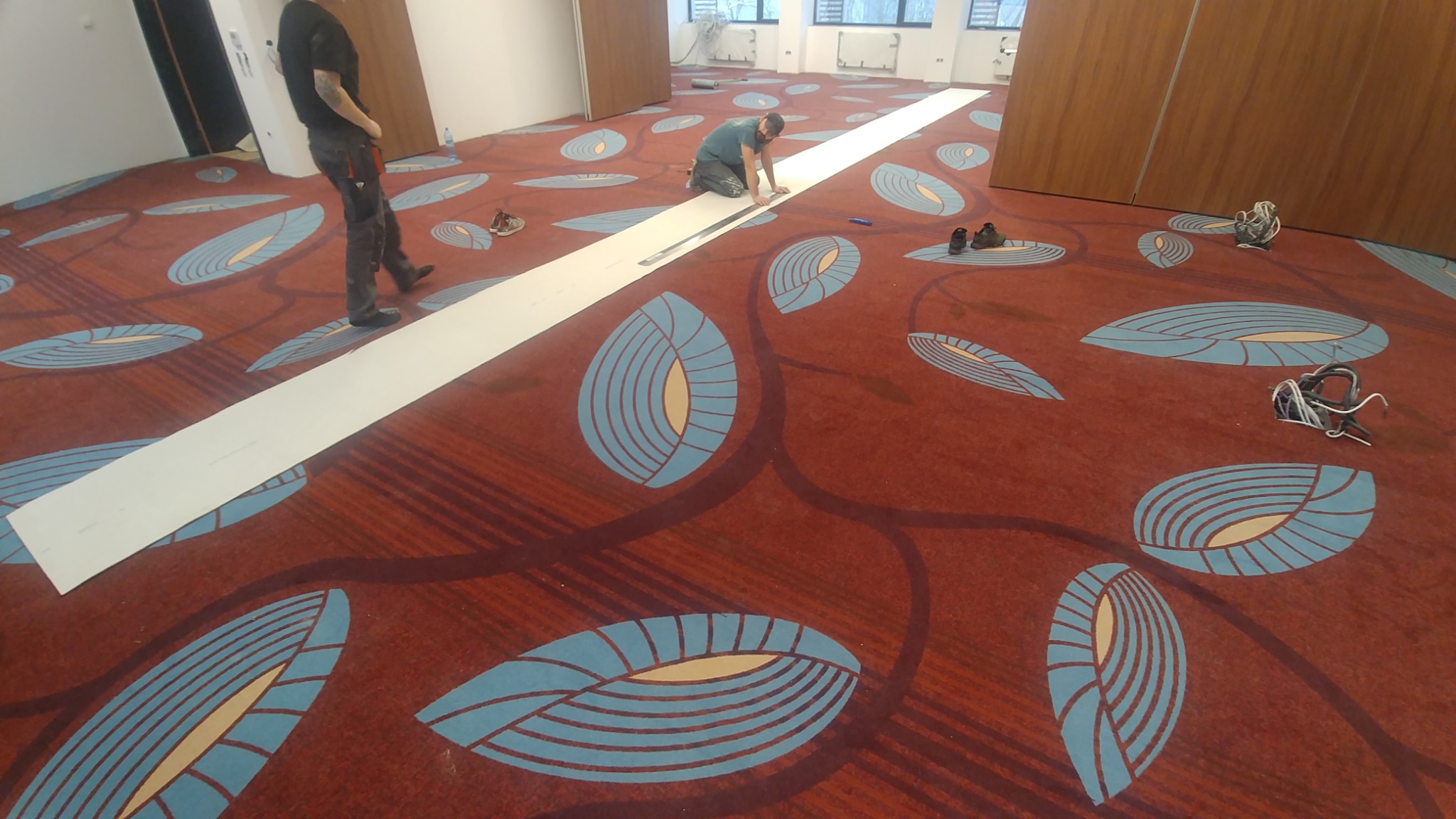 Pracownik mierzy wykładzinę wzorzystą w odcieniach czerwieni i błękitu w przestronnym pomieszczeniu, przygotowując ją do przycięcia wzdłuż długiej listwy pomiarowej.