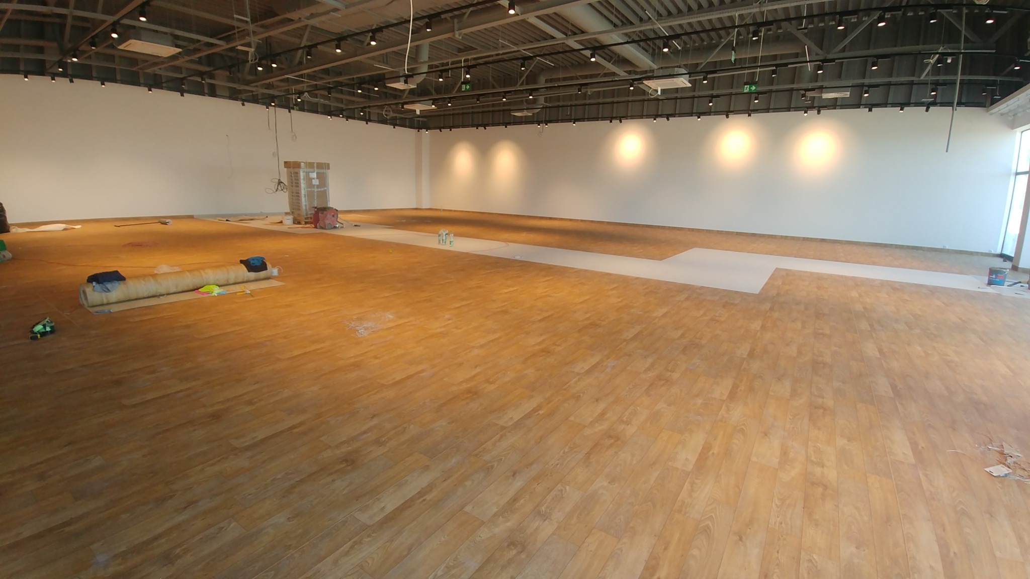 Duża, pusta przestrzeń komercyjna w trakcie układania podłogi z paneli imitujących drewno, z widocznym rozwiniętym rolką materiału podkładowego i częściowo ułożonymi panelami.