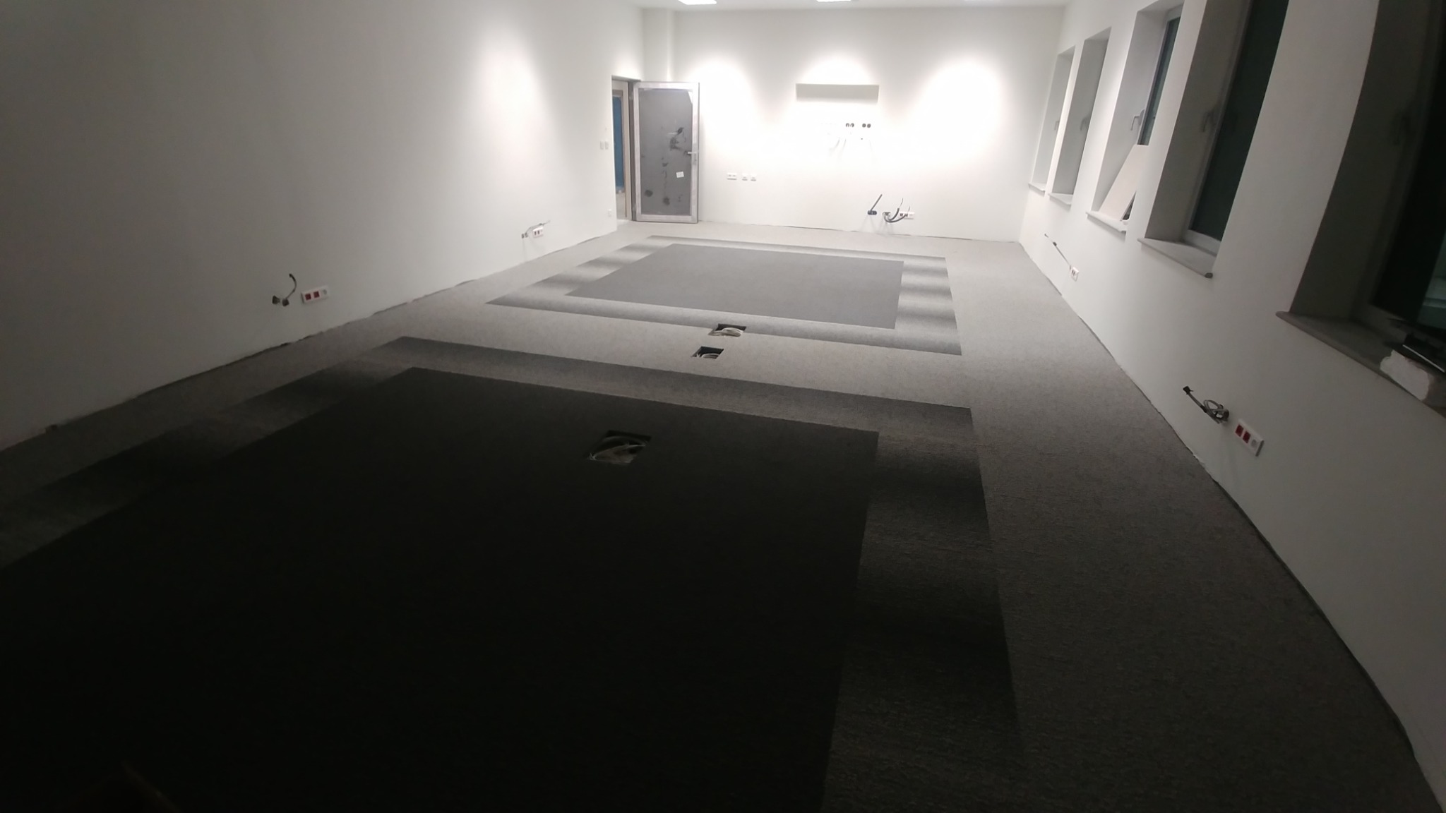 Układana szara wykładzina dywanowa w pustym, jasnym pomieszczeniu biurowym z widocznymi puszkami podłogowymi i oknami.
