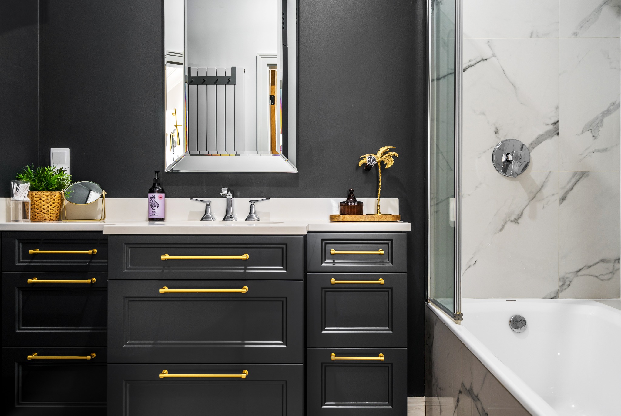 Elegancka, czarno-biała łazienka w Warszawie z ciemnymi szafkami z złotymi uchwytami, marmurowymi płytkami i wanną za szklanym parawanem.