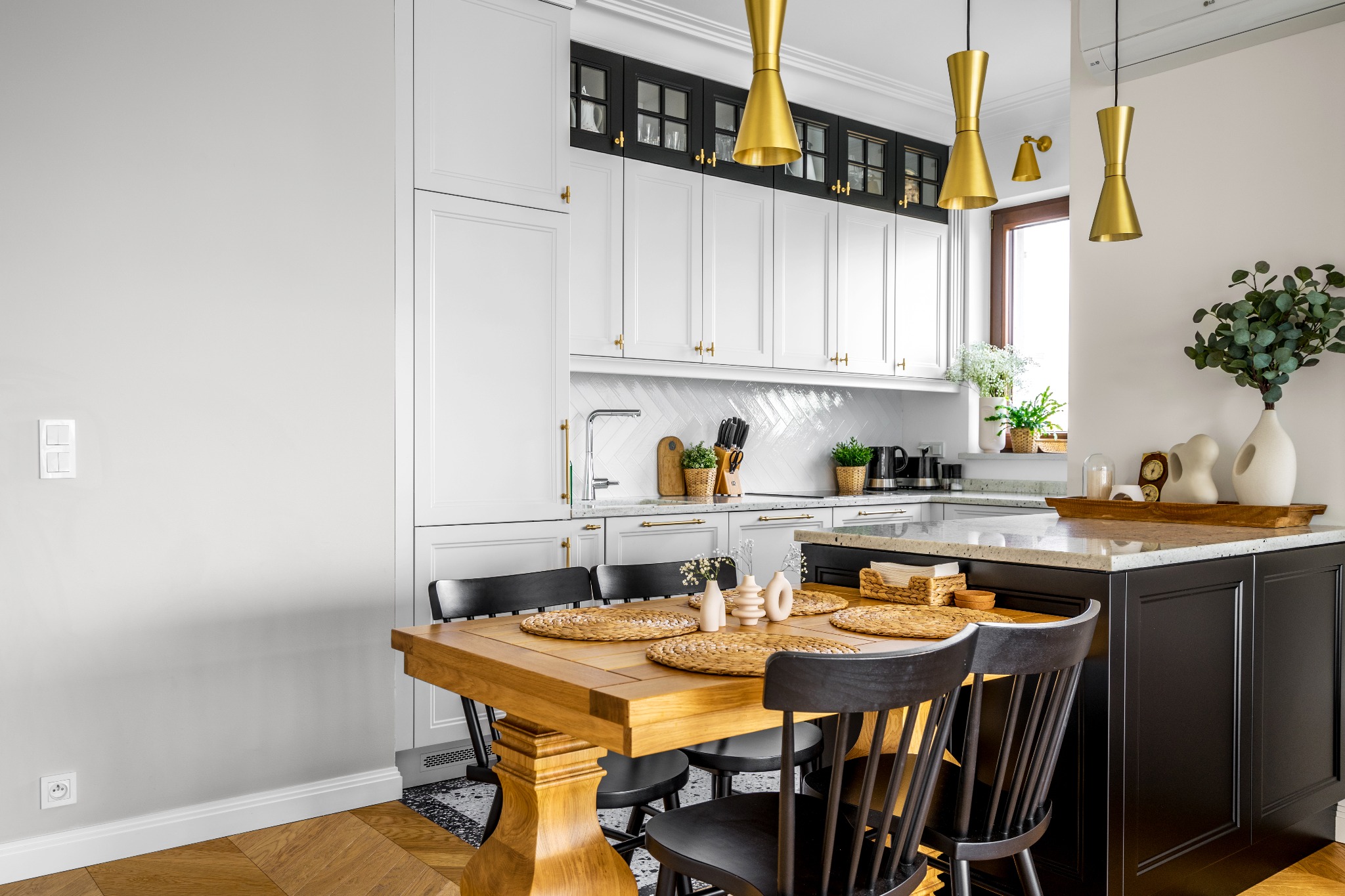 Jasna kuchnia z drewnianym stołem i czarnymi krzesłami, złote lampy wiszące nad wyspą, białe szafki z czarnymi akcentami i złotymi uchwytami, parkiet w jodełkę na podłodze.