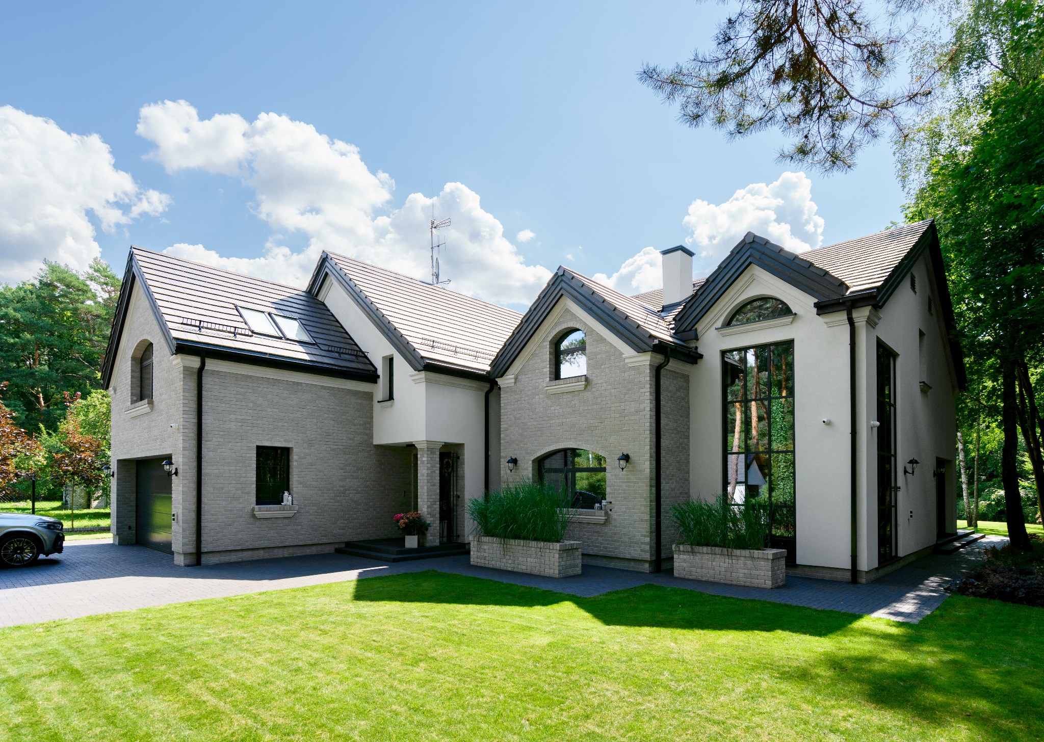 Elegancki, dwukondygnacyjny dom z szarej cegły z ciemną dachówką, dużymi oknami i zadbanym trawnikiem przed budynkiem w słoneczny dzień.