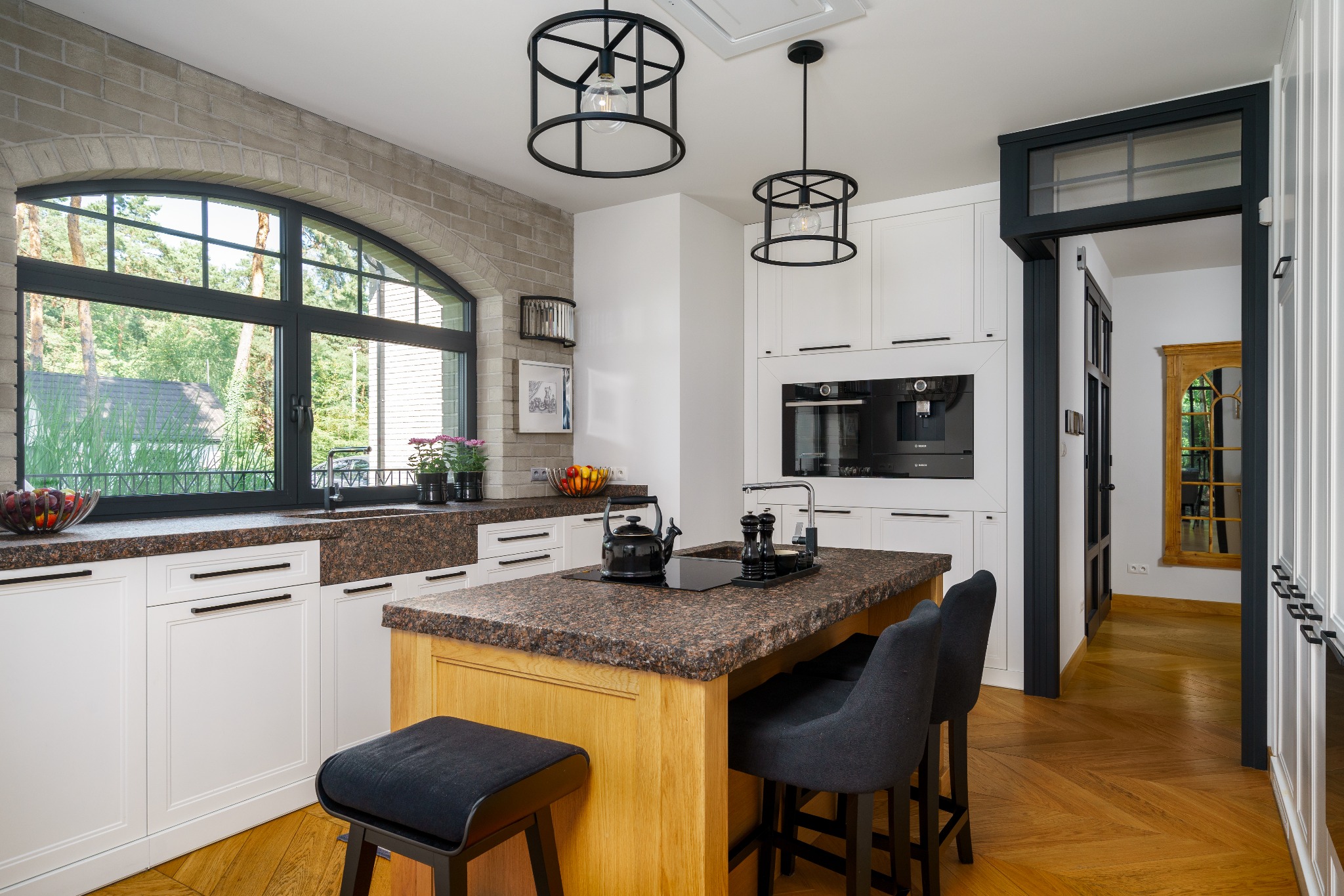 Nowoczesna kuchnia z wyspą, brązowym blatem i czarnymi detalami, okrągłe lampy wiszące, duże okno z widokiem na zieleń.