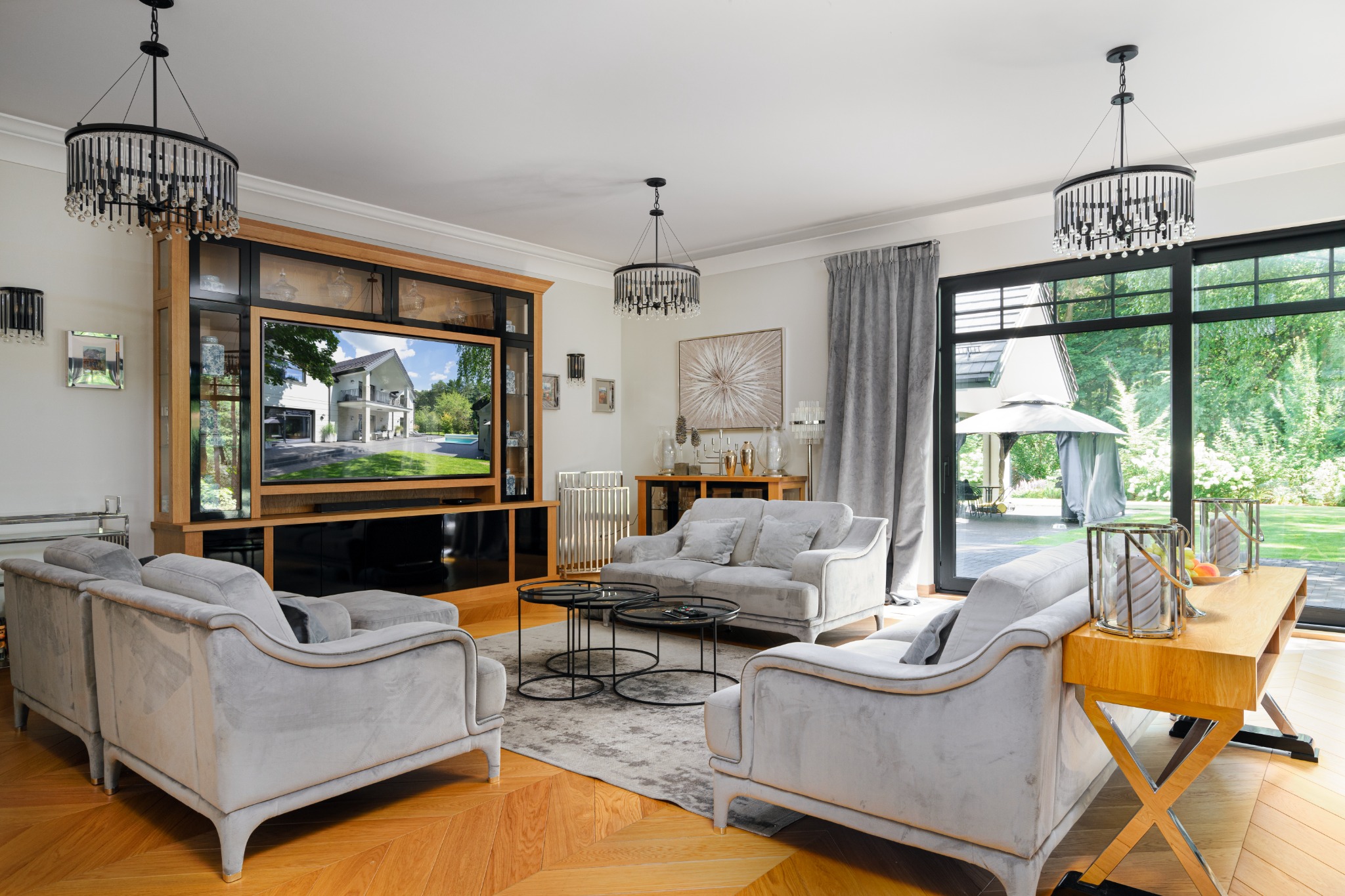 Elegancki salon z szarymi sofami i parkietem w jodełkę, oświetlony wiszącymi żyrandolami, z widokiem na ogród przez duże okno.