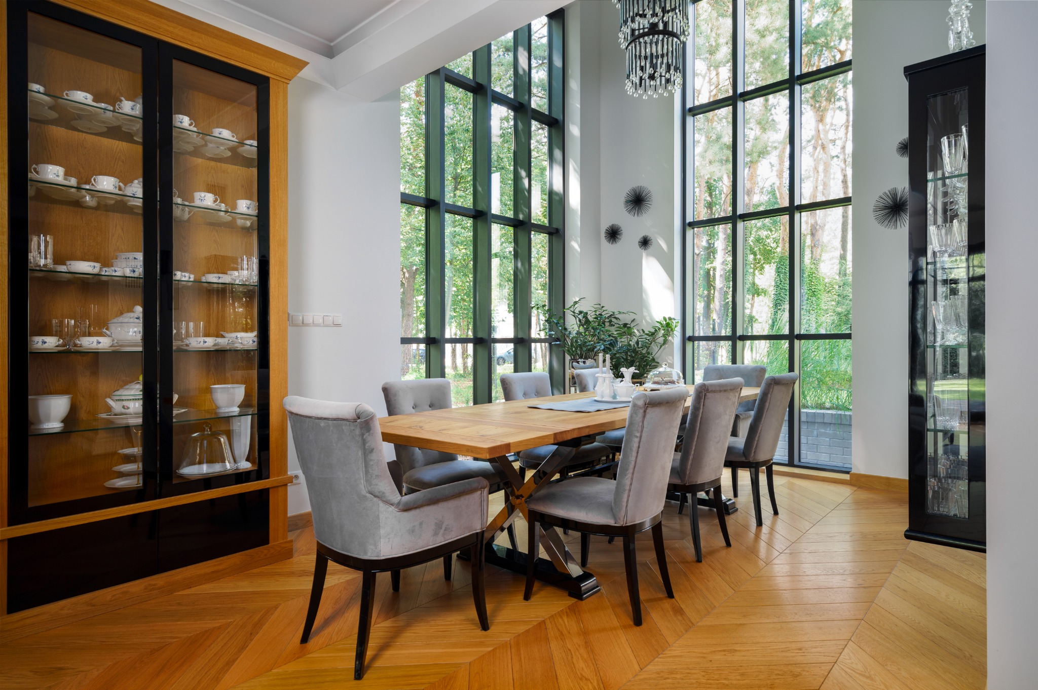 Jasna jadalnia z parkietem w jodełkę, dużym oknem z widokiem na zieleń, drewnianym stołem z szarymi krzesłami i dwoma przeszklonymi witrynami z zastawą.